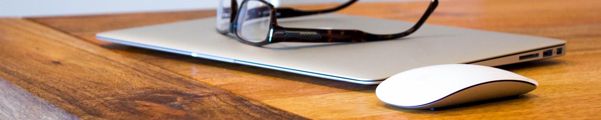 Houten tafel met dichtgeklapte laptop, bril en computermuis.
